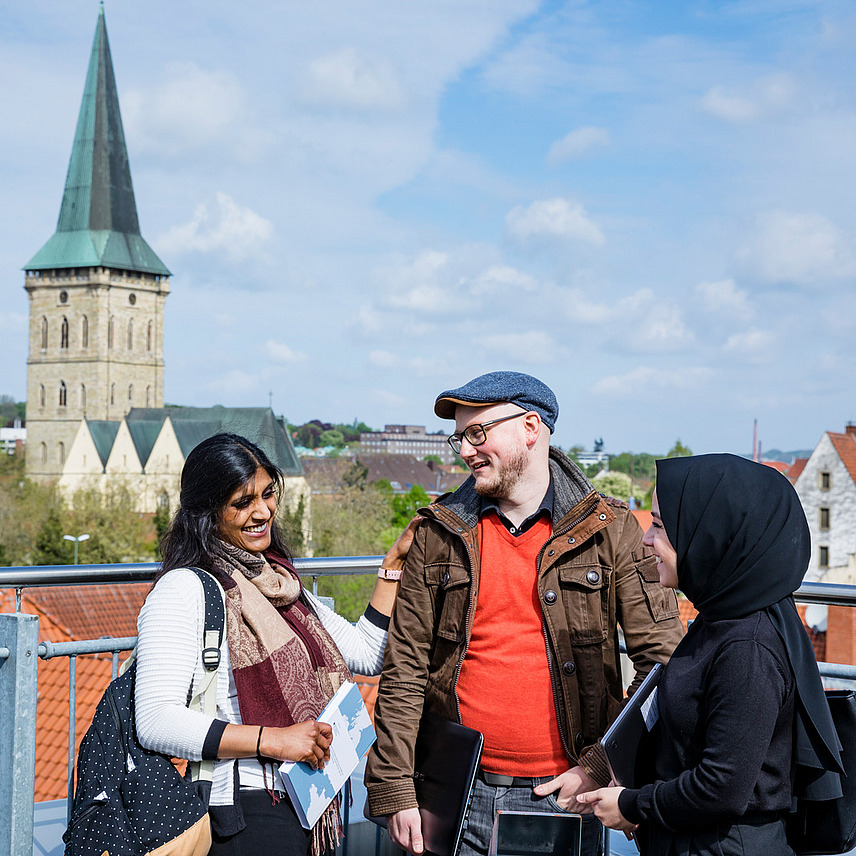 Three people stand in front of a railing with a view over the rooftops of Osnabrück. One of them is wearing a headscarf.