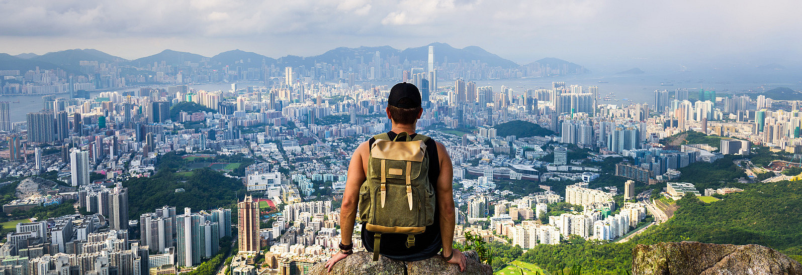 A young man stands on a mountain with a rucksack and looks at the town in the valley in front of him, right by the sea.