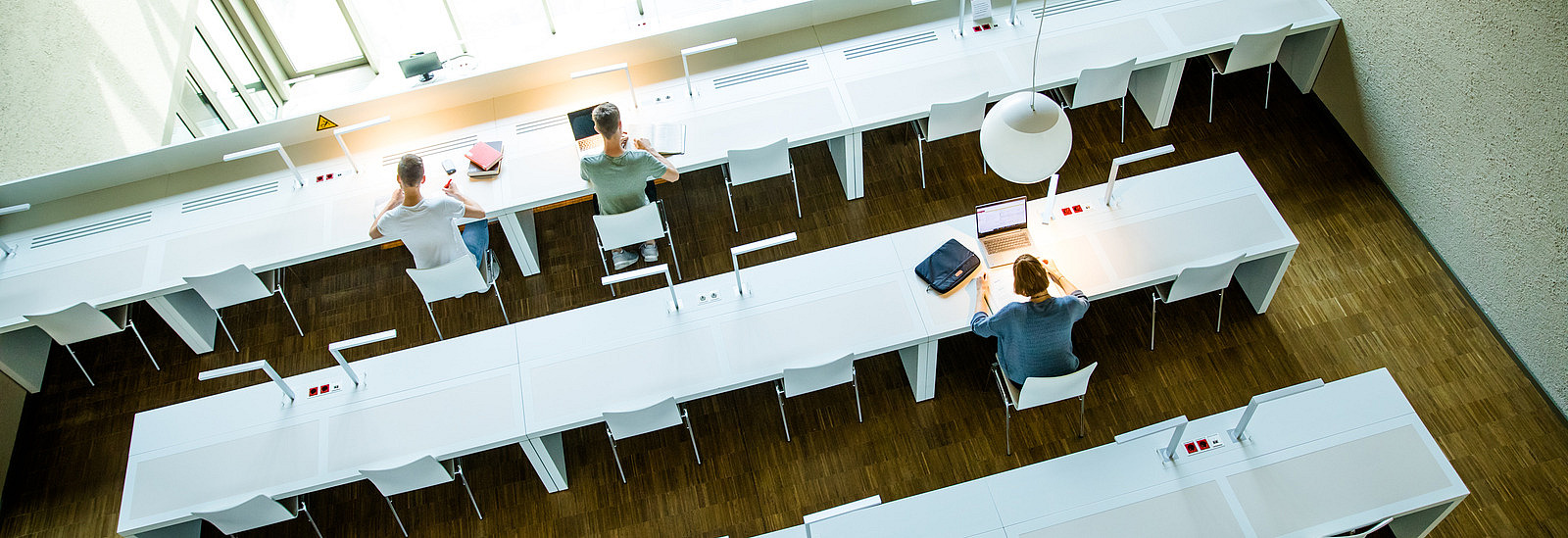 View from above into a modern reading room with long white tables and chairs. Three people are sitting at different tables, working on their laptops or reading. Large windows let in plenty of daylight and practical lamps provide additional light. The floor is made of wood.