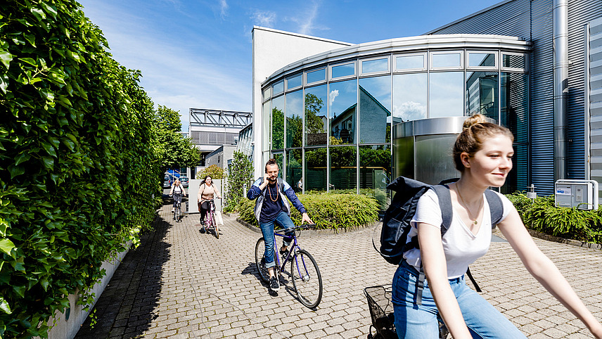 Four students ride past a building with a glass and steel façade on bicycles.