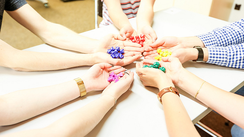 Five people form a circle with their hands in the middle of a table. In each of their hands are different colored dice.