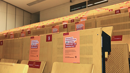 In a lecture hall posters are hung up showing the slogan "Sexualized discrimination and violence - [Ir]relevant for universities?!" and further information