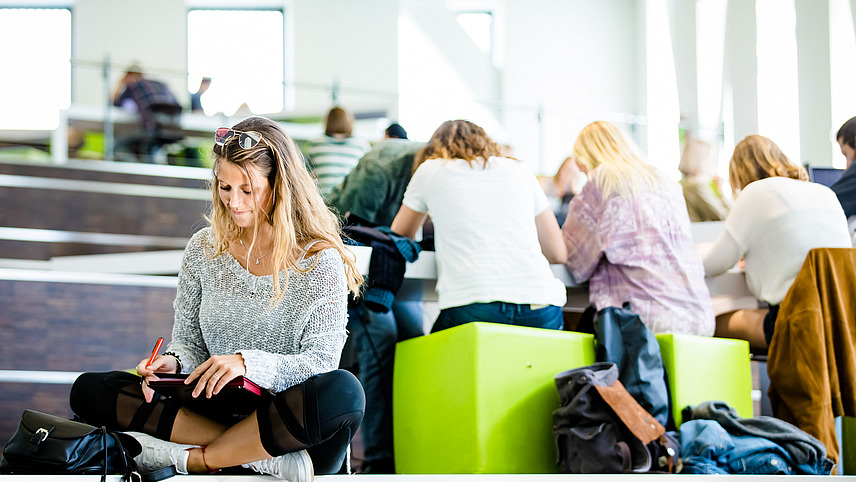 Studentin sitzt im Schneidersitz und schreibt, im Hintergrund sind weitere Studierende zu sehen