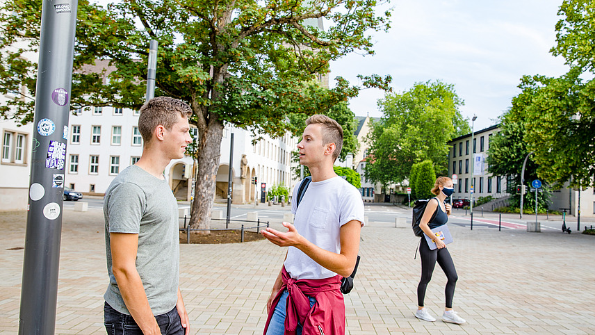 Zwei Studenten unterhalten sich auf einem bepflasterten Platz mit vereinzelten Bäumen. Eine Studentin geht mit ihrem Laptop und aufgesetzer medizinischer Maske an ihnen vorbei.