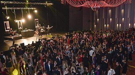 View from an elevated position over the guests at the university ball. A band with a singer, backing vocalist, guitar, bass guitar and drums is playing on stage. Above the dance floor hangs a huge chandelier made of shining red hanging pearl necklaces and a disco ball in the middle.