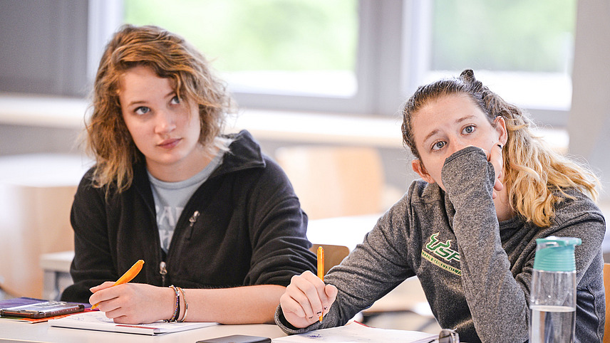 Eine rothaarige und eine blonde Frau sitzen in einem Kursraum und machen sich Notizen während sie aufmerksam nach Vorne schauen.