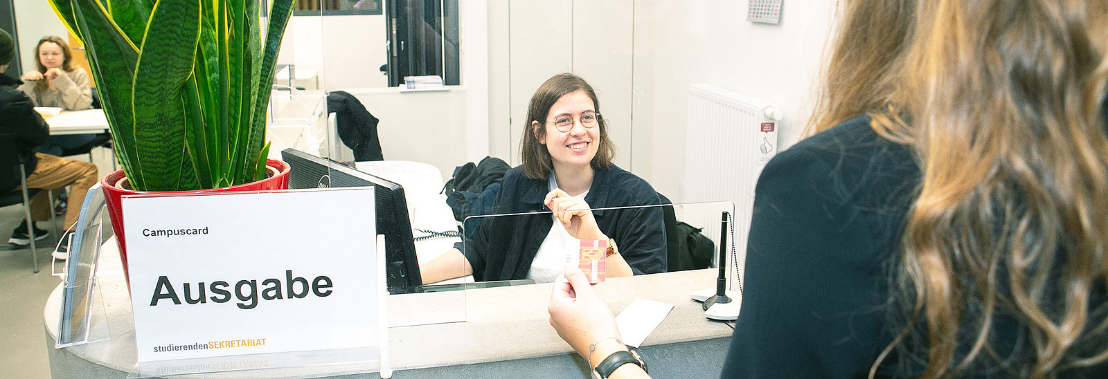 A staff member at the InfoPoint is smiling and holding a campus card. In the background, a few students can be seen at tables, and a large plant is on the reception desk.