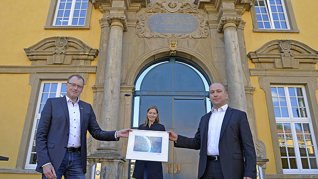 Two men and a woman stand in front of a historic yellow façade with a large entrance door, while one of the men holds a picture in his hands.