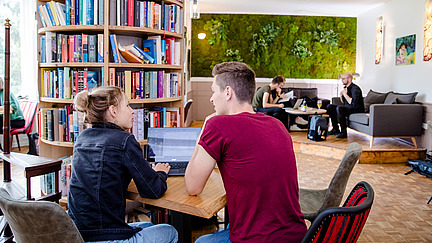 Two students are sitting in a café next to a round bookshelf and talking. In front of them is a laptop.