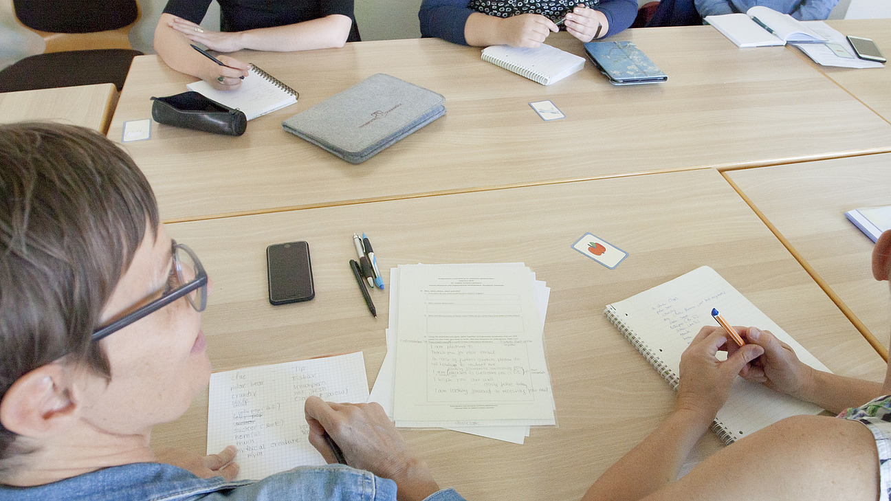 A table with several people attending a meeting. They are taking notes. Writing materials such as notepads, pens and a laptop are on the table.