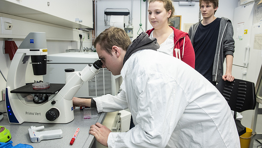 A man stands in a laboratory looking through a microscope at a Petri dish with red contents. Two other people stand behind him and observe the process.