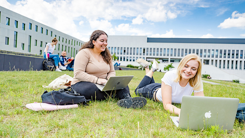 Zwei Studentinnen auf dem begrünten Platz neben der Bibliothek. Eine von ihnen sitzt, die andere liegt auf dem Rasen, beide haben Laptops vor sich