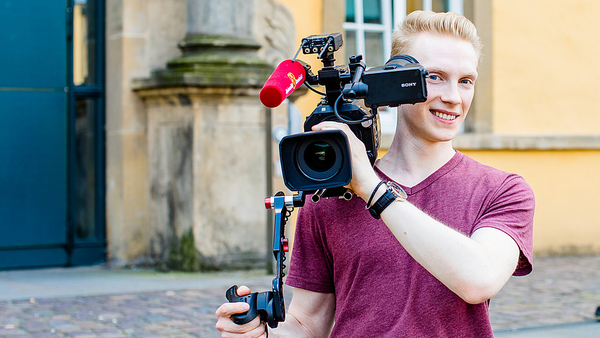  A young man holds a camera ready to film in front of Osnabrück University's castle building. 