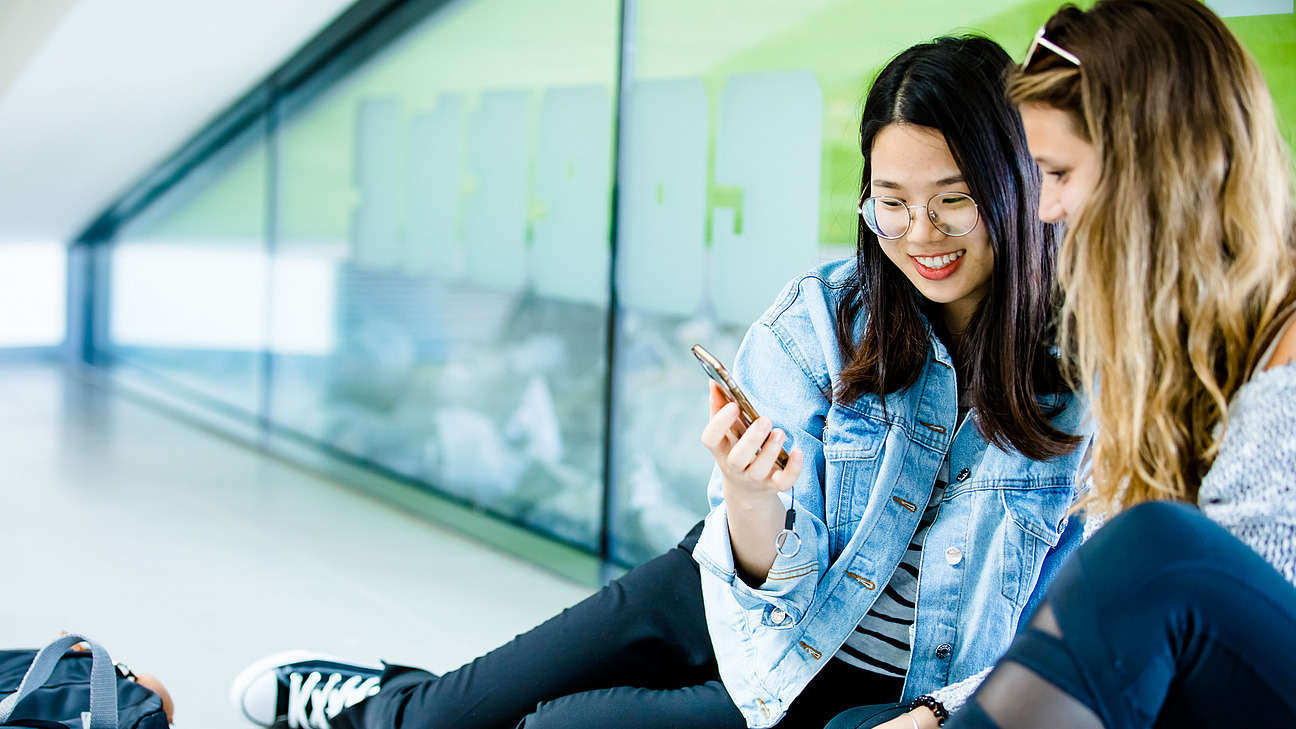 Zwei junge Frauen sitzen auf dem Boden und schauen fröhlich auf ein Smartphone