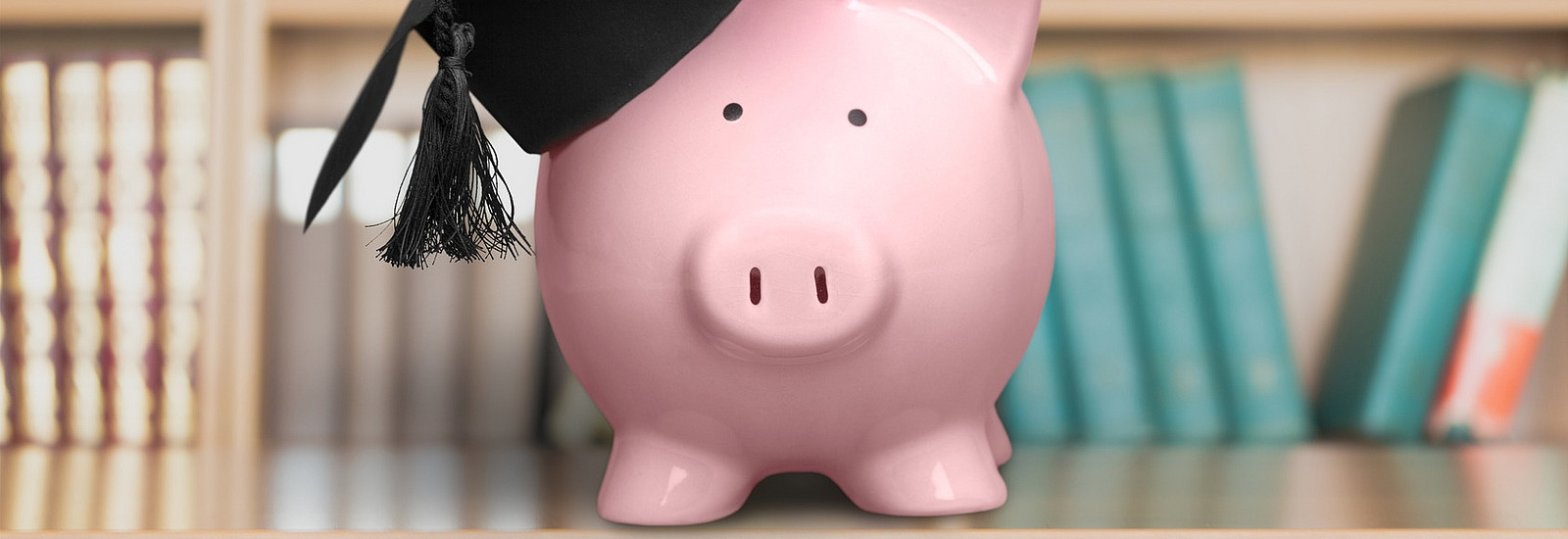 A pink piggy bank wearing a black graduation cap. In the background, there are bookshelves filled with books of various colors.