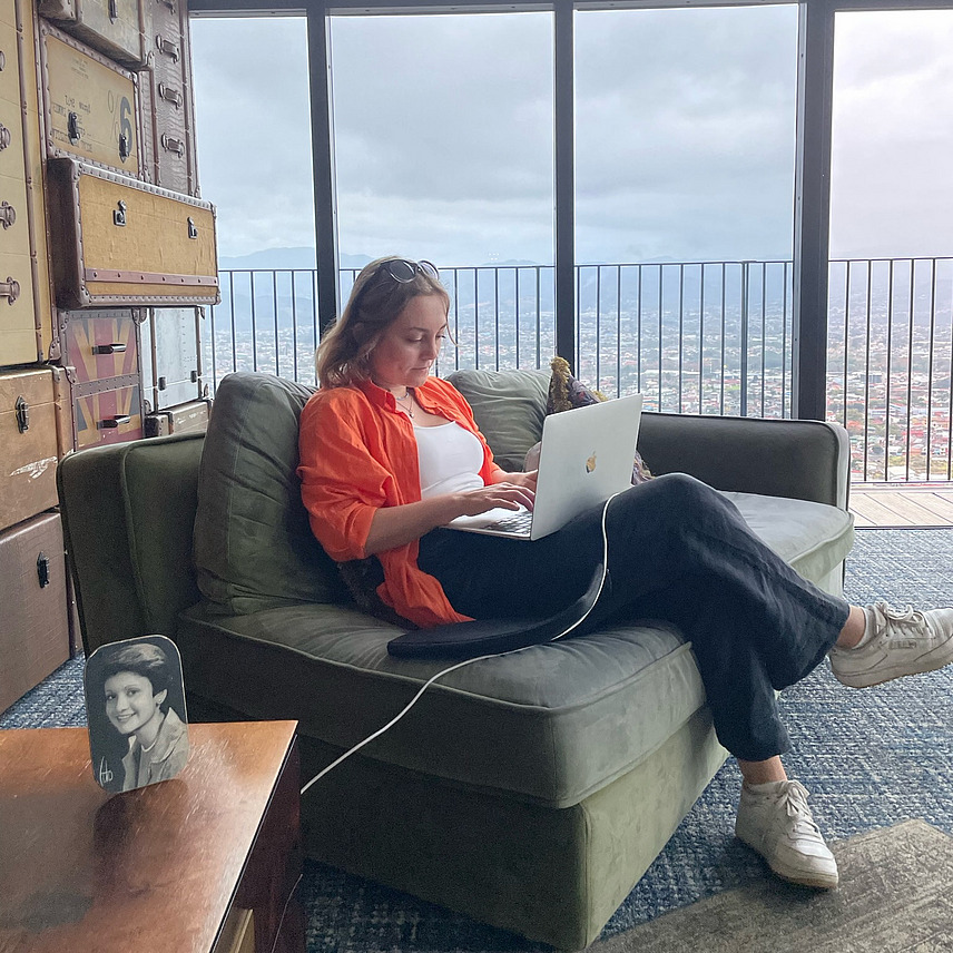 Dilay Dollnick sits on a sofa and works on her PC. You can see the Costa Rican landscape through the large windows to her left.