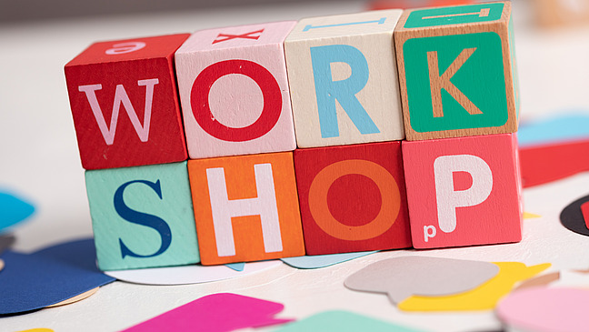 Wooden blocks that form the word "WORKSHOP". The blocks are designed in different colors, including red, blue, pink, green and beige. They are surrounded by various colored paper shapes and fragments.