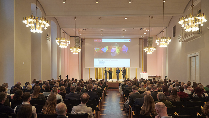 A choir sings on stage in a large hall. Many people are sitting in the audience.