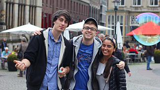 Three international students stand smiling on a market square in the center of Bremen.