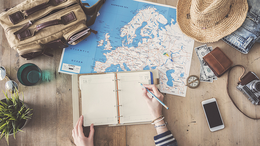 Symbolic image for preparing a trip. Hat, mobile phone, camera, wallet and a bag lie next to a map of Europe and a person makes notes in a small diary.