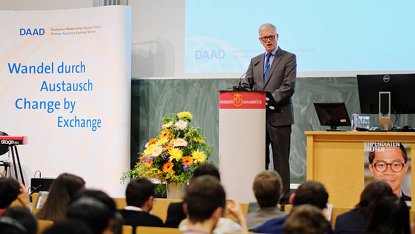 DAAD event at Osnabrück University, where an elderly man gives a speech in front of an audience and a DAAD presentation is projected on the wall in the background.