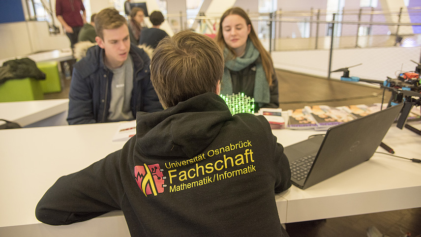 A student wearing a hoodie is standing at a table and talking to two other students. On the table, there is a laptop and some brochures. In the background, there is an open and bright space with more people and furniture.