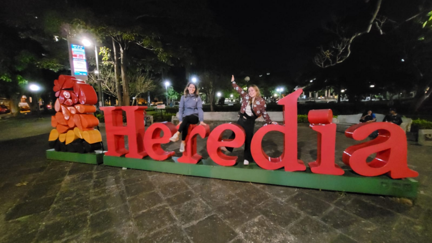 The image shows a brightly lit scene at night featuring a large sign "Heredia" in bold red letters. Two people are sitting on / near the sign. The background includes trees and decorative lights.