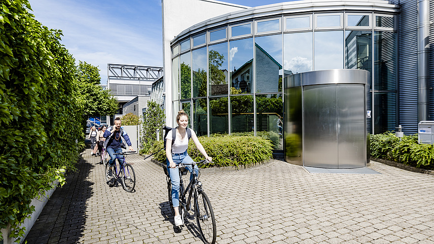 Four students ride past a building with a glass and steel facade on bicycles in sunny weather.