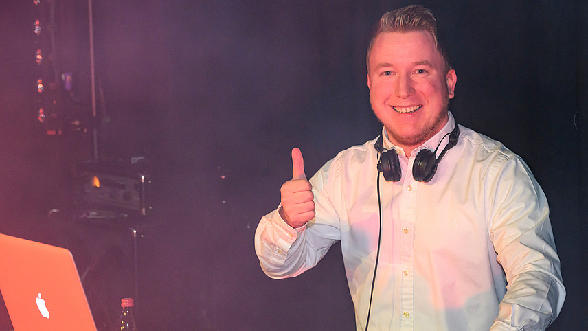 A person in front of a mixing desk smiles at the camera and gives a thumbs up