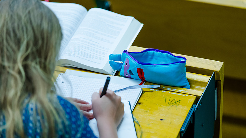 A student makes a note in her planner while sitting on a yellow bench in a lecture hall.