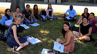 A group of international students sit in a circle on the grass and work together in the summer temperatures.