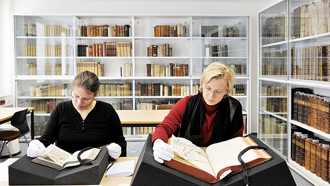 Das Bild zeigt zwei Frauen, die in einem weißen, modernen Raum alte Bücher studieren. Beide tragen weiße Handschuhe und arbeiten an einem Tisch mit speziellen Buchstützen, um die Bücher zu halten. Die Frau auf der linken Seite liest in einem kleinen Buch, während die Frau auf der rechten Seite sich auf ein größeres, geöffnetes Buch konzentriert