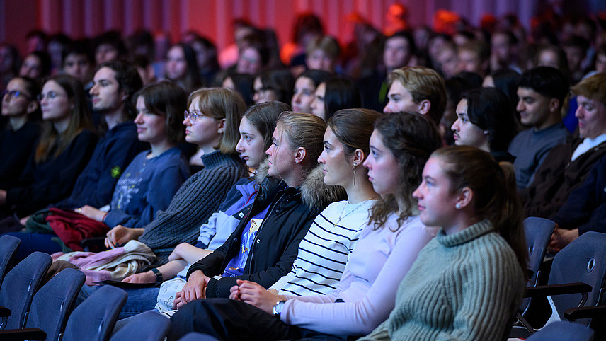 Nahaufnahme von gefüllten Zuschauerreihen in einem Saal. Die Personen blicken interessiert Richtung Bühne.
