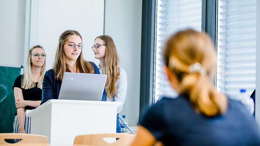 Drei Studentinnen stehen in einem Seminarraum vor einem Podium. Eine von ihnen schaut auf einen Laptop und spricht, während das Publikum zuhört.