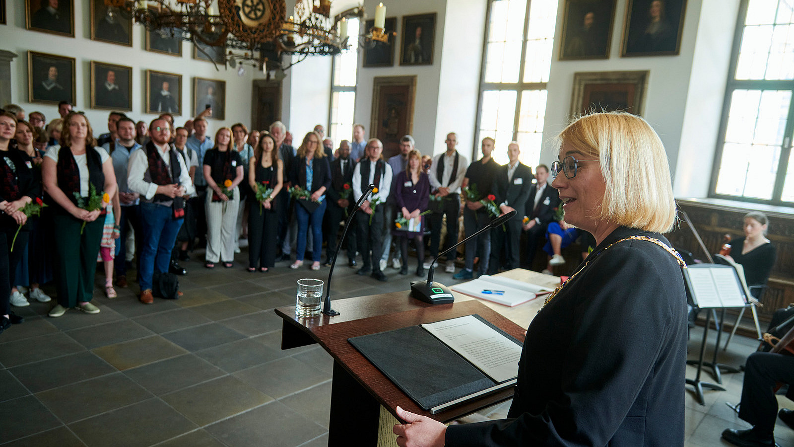 In einem historisch wirkendem Raum steht im Vordergrund eine Person an einem Rednerpult. Vor ihr stehen zahlreiche Personenn und schauen sie an.