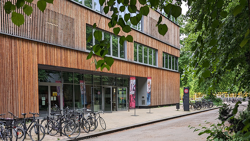 Ein modernes Gebäude mit Holzfassade und großen Fenstern, umgeben von Bäumen. Vor dem Gebäude stehen mehrere Fahrräder abgestellt, und die Eingänge sind sichtbar. Die Straße führt am Gebäude vorbei.