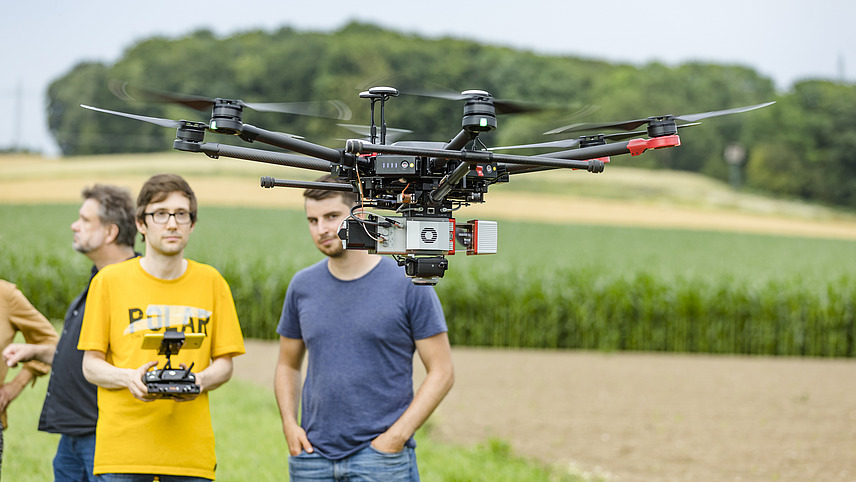 Drone hovers in the air. A few people are standing around it.