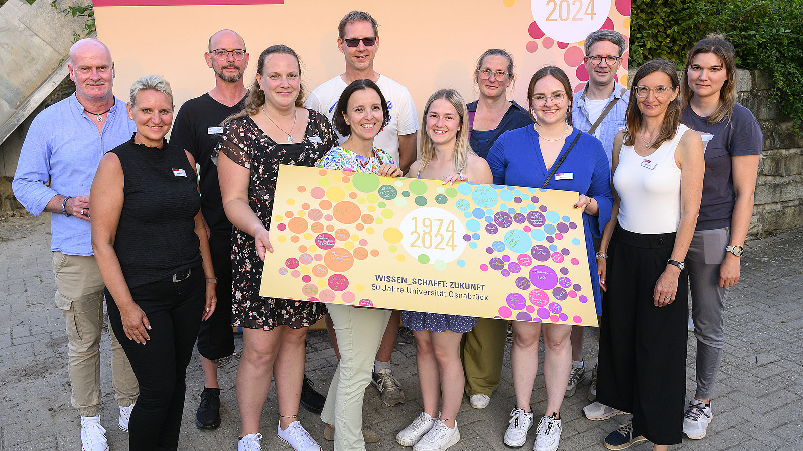 A group of people stands in front of a photo background and holds a large colorful sign into the camera.