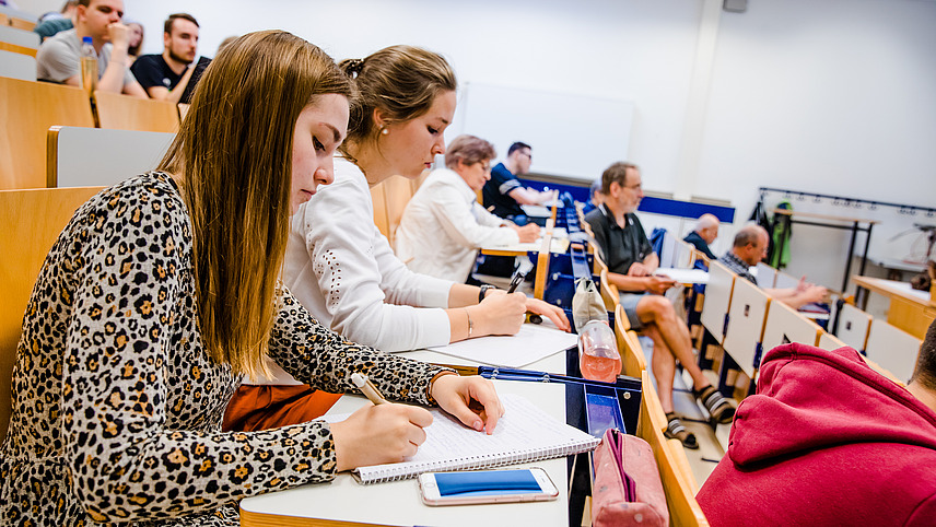 Zwei Studentinnen in einer Sitzreihe in einem Vorlesungssaal. Sie machen sich Notizen. Im Hintergrund sind weitere Studierende auf den gelben Hörsaalbänken zu sehen.