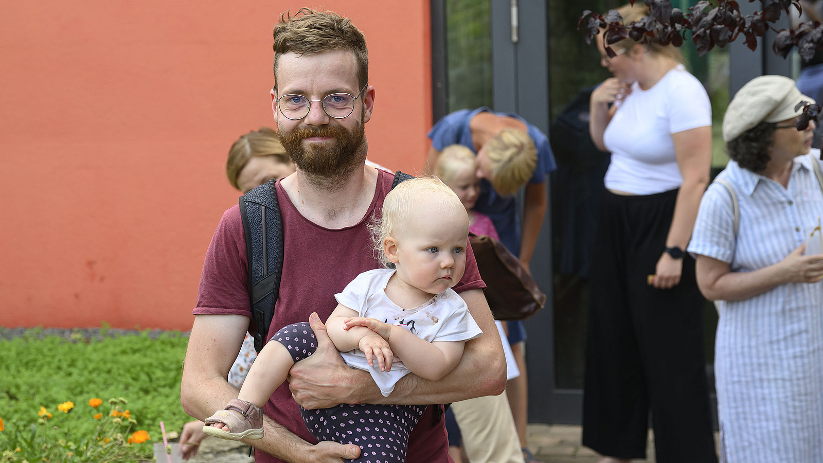 A man carries a toddler in his arms and smiles at the camera.