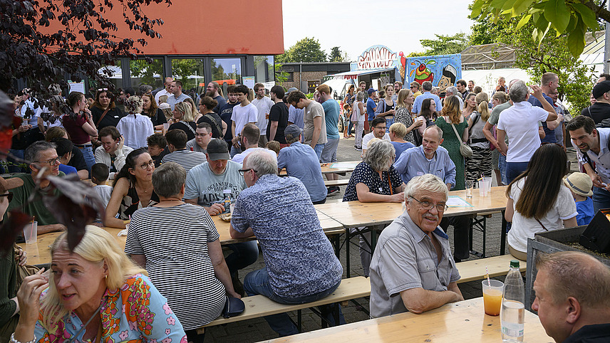 Zahlreiche Menschen sitzen auf Bänken an Tischen bei einer Party.