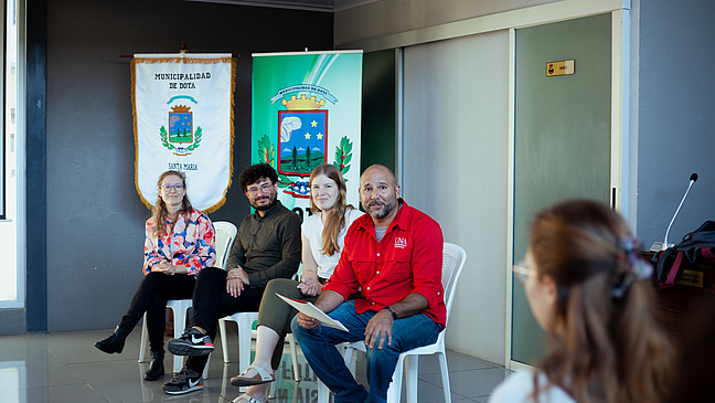 Eine Podiumsdiskussion in einem Raum. Vier Personen sitzen auf Stühlen, im Hintergrund sind zwei Banner mit Symbolen und Text zu sehen. Die Person ganz rechts hält ein Blatt Papier in der Hand. Im Vordergrund sind teilweise Zuhörer sichtbar. 