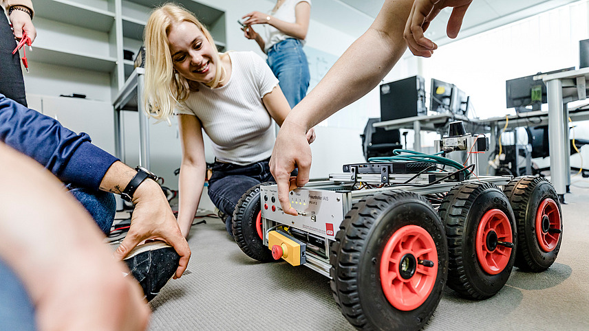 A few students kneel in front of a box with wheels. Many cables are sticking out of the box and there are many buttons and switches on one side. One of the students presses one of the switches.