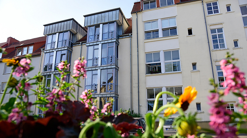 Im Vordergrund ein Blumenbeet und im Hintergrund ein Wohnhaus mit Fensterfront und links daneben ein Wohnhaus mit verglasten Erkern.