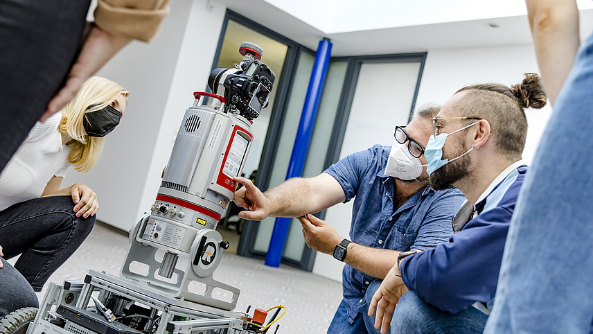 Ein Mann kniet zusammen mit ein paar Studierenden vor einem Gerät mit Rädern und einer Kamera an einem mechanischen Arm. Er zeigt den Studierenden etwas am Arm des Gerätes. Alle Personen tragen eine medizinische Maske.