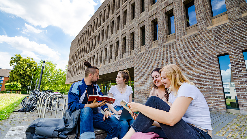 Vier Studierende sitzen vor der Bibliothek am Westerberg auf einer Bank und unterhalten sich, sie halten Bücher in der Hand, eine Studentin hat einen Laptop