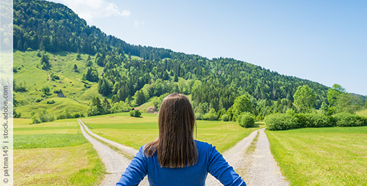 Eine Frau steht mit dem Rücken zur Kamera an einem Weg der sich Gabelt