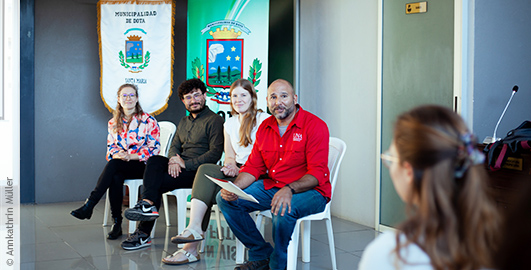 Das Bild zeigt vier Personen, die in einem Konferenz- oder Besprechungsraum sitzen. Sie sind in einem Halbkreis angeordnet. Hinter ihnen befinden sich Fahnen oder Banner, die die "Municipalidad de Dota" repräsentieren und ein Wappen oder Emblem zeigen. Die Personen scheinen in eine Diskussion vertieft zu sein. Der Raum wirkt modern und hell, mit gefliesten Böden.