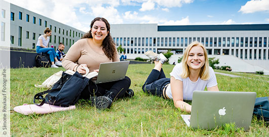 Zwei Studentinnen auf dem begrünten Platz neben der Bibliothek. Eine von ihnen sitzt, die andere liegt auf dem Rasen, beide haben Laptops vor sich