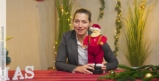 Eine Frau vor festlich geschmücktem Hintergrund hält eine Weihnachtsmannpuppe im Arm